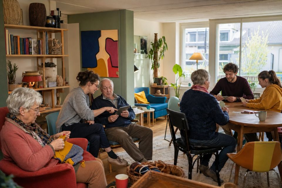 Unverbindliche KI-Konzeptvisualisierung eines wohnlichen Treffpunkts mit älteren und jüngeren Nachbarn bei Handarbeit, Kartenspiel und Tablet-Hilfe.
