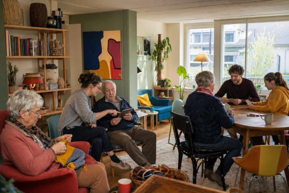 Unverbindliche KI-Konzeptvisualisierung eines wohnlichen Treffpunkts mit älteren und jüngeren Nachbarn bei Handarbeit, Kartenspiel und Tablet-Hilfe.