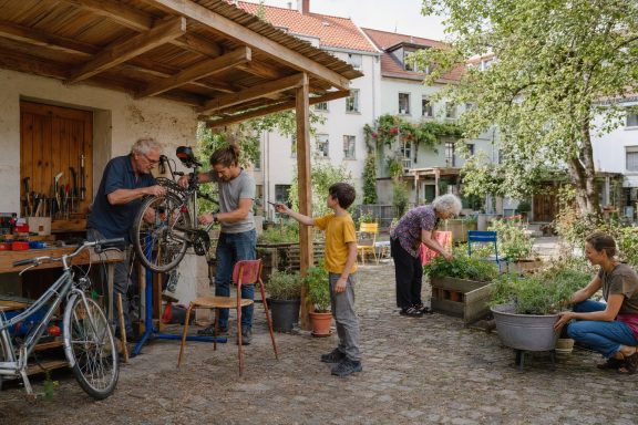 Unverbindliche KI-Konzeptvisualisierung einer offenen Hofwerkstatt mit Fahrradreparatur, Hochbeeten und generationenübergreifender Alltagshilfe.